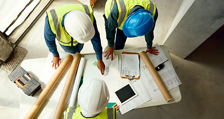 an overhead shot of three construction workers wearing hardhats and hi-vis jackets reviewing plans